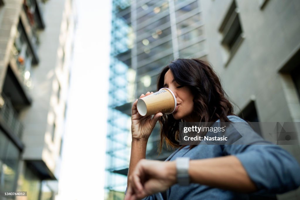 Junge Geschäftsfrau, die ihre Armbanduhr vor dem Bürogebäude anschaut und Kaffee trinkt