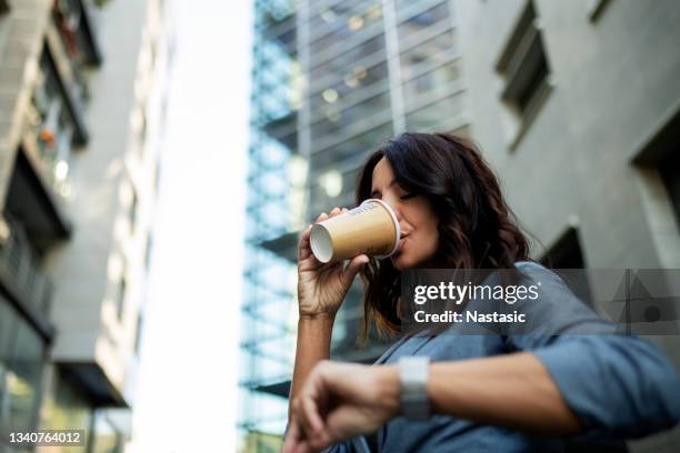 junge geschäftsfrau, die ihre armbanduhr vor dem bürogebäude anschaut und kaffee trinkt - wartezeit stock-fotos und bilder