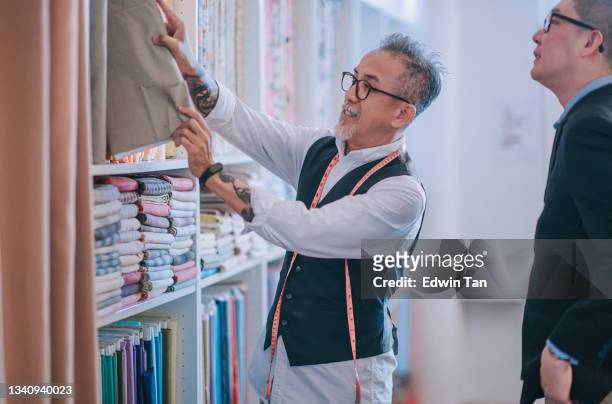 asian chinese senior man tailor picking and showing his customer on fabric garment choice on new tailored clothing suggestion at his store - craft show stock pictures, royalty-free photos & images