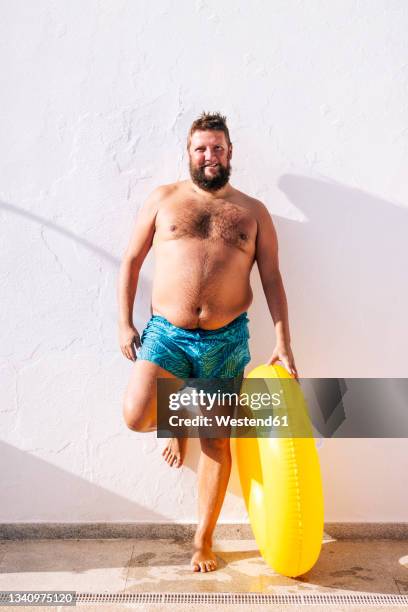 shirtless man with inflatable ring leaning on wall during sunny day - chubby swimsuit stock pictures, royalty-free photos & images