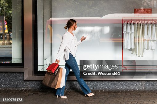 Young woman using smart phone while walking with shopping bags by store Young woman using smart phone while walking with shopping bags by store