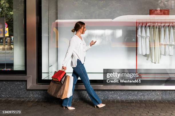 young woman using smart phone while walking with shopping bags by store - bärkasse bildbanksfoton och bilder