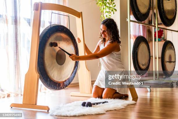 female sound therapist playing gong while sitting on stool in studio - gong fotografías e imágenes de stock