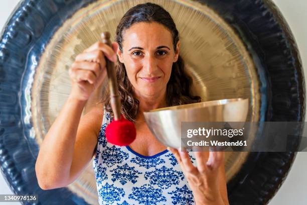 smiling female sound therapist holding singing bowl and mallet in studio - percussion mallet stock-fotos und bilder