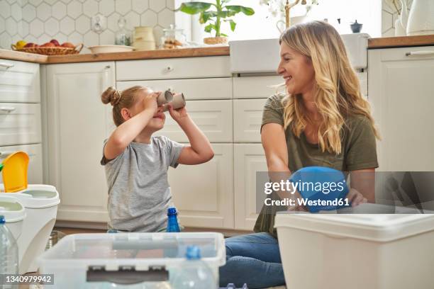 playful girl looking at mother through rolled up paper in kitchen - recylcebak stockfoto's en -beelden