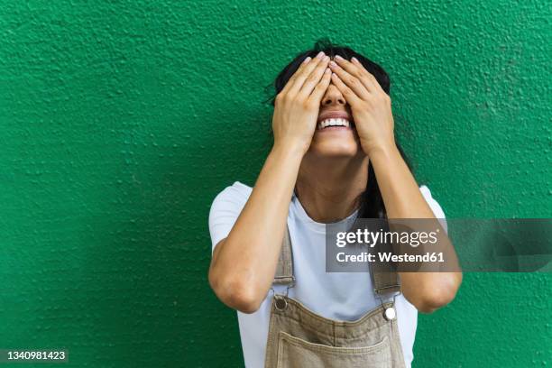 smiling young woman covering eyes with hands in front of green wall - hands covering eyes stock pictures, royalty-free photos & images