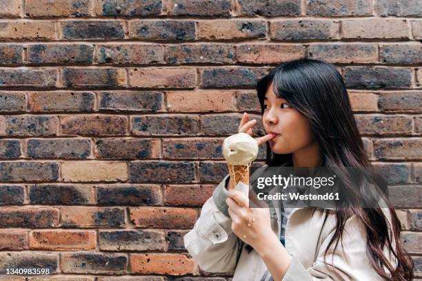 woman licking finger while holding ice cream in front of brick wall - woman ice cream stock pictures, royalty-free photos & images