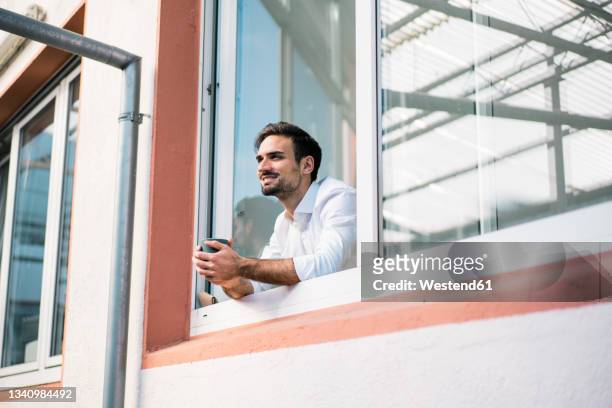 smiling young male professional holding coffee cup while standing at window during break - looking outside window stock-fotos und bilder