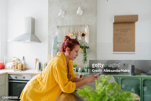 mature woman using smart phone in a kitchen of her apartment - vrouw 50 jaar stockfoto's en -beelden