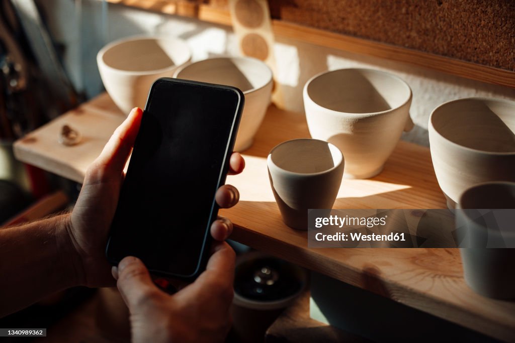 Male artist using smart phone by clay pots on table at home office