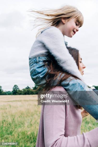 mother carrying smiling daughter while playing on meadow - piggyback stock pictures, royalty-free photos & images