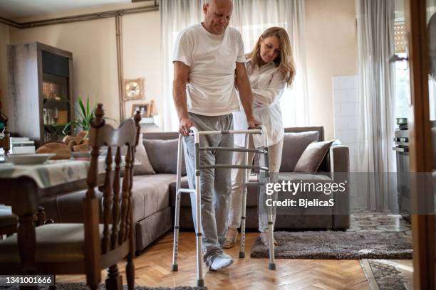wife helping her injured husband to walk using walking frame at home - walking frame stock pictures, royalty-free photos & images