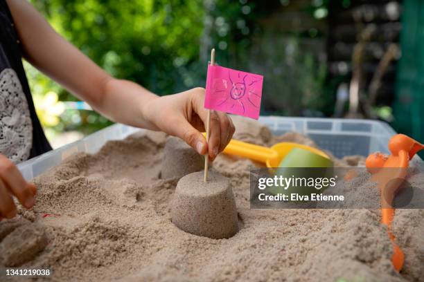 child placing a homemade flag made from a pink sticky note with a drawing of a smiley face, into the top of a small sandcastle surrounded by sand inside a plastic sandbox. - zandbak stockfoto's en -beelden