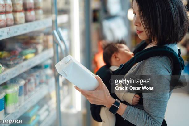 young mother doing grocery shopping with her baby daughter in a carrier at supermarket - mom buying milk stock pictures, royalty-free photos & images