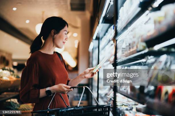 young asian woman carrying a shopping basket while grocery shopping in supermarket, standing along the dairy aisle, reading the nutrition label on a packet of fresh organic cheese. making healthier food choices - loja de conveniencia imagens e fotografias de stock