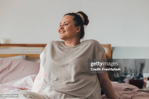 portrait d’une belle femme de taille plus assise sur un lit dans sa chambre - mannequin grande taille photos et images de collection