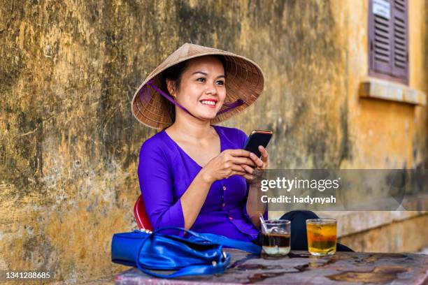 vietnamese woman using mobile phone during coffee break, old town in hoi an city, vietnam - ao dai stockfoto's en -beelden