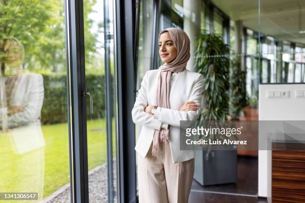muslim businesswoman standing in office and looking outside - femme foulard photos et images de collection