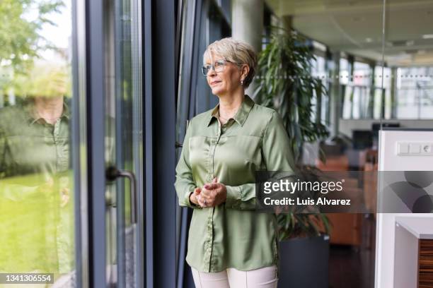 senior woman standing by a window in office - dreiviertelansicht stock-fotos und bilder