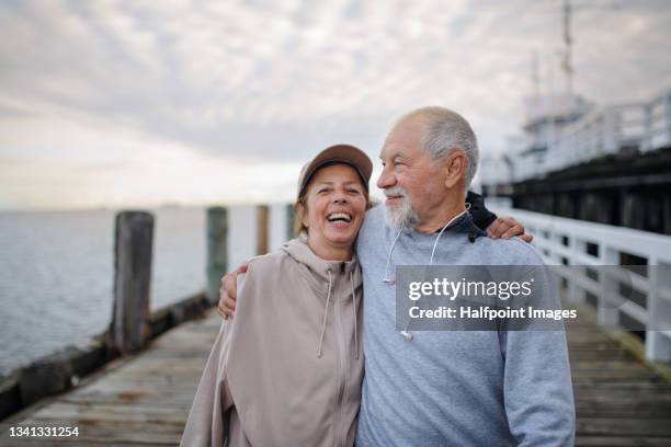 active senior couple runners taking break during doing exercise outside on the pier by the sea, looking at camera. - senior adult couple stock pictures, royalty-free photos & images