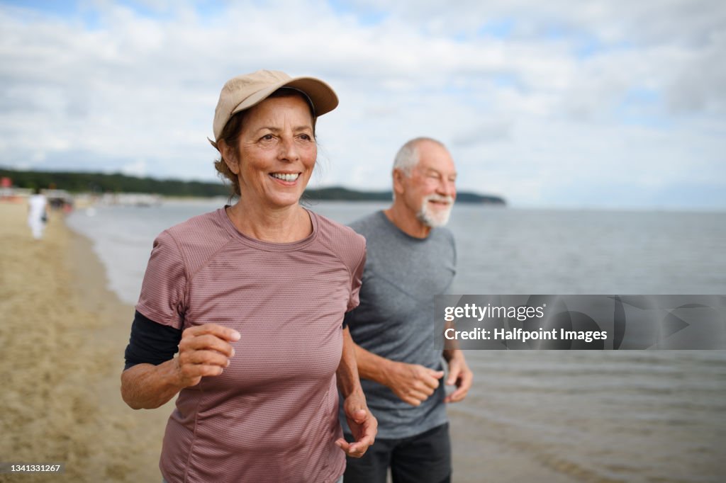 Active senior couple runners jogging outdoors on sandy beach by sea in early morning.