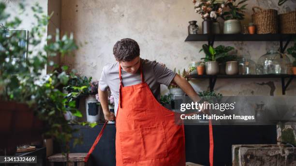 young male florist with down syndrome putting on apron indoors in flower shop. - apron stockfoto's en -beelden