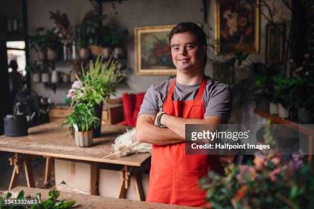 young male florist with down syndrome standing indoors in flower shop, looking at camera and smiling. - three quarter length stockfoto's en -beelden
