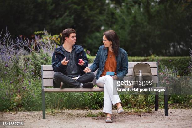 young down syndrome man with his mother resting and having snack outdoors in park. - park bench fotografías e imágenes de stock