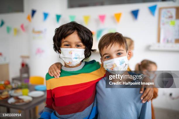two classmates boys with face masks standing and holding sholuders inside in classrom at school, coronavirus concept. - togetherness covid stock pictures, royalty-free photos & images