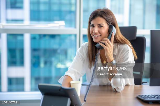 businesswoman using landline and smiling at her desk - landline phone imagens e fotografias de stock