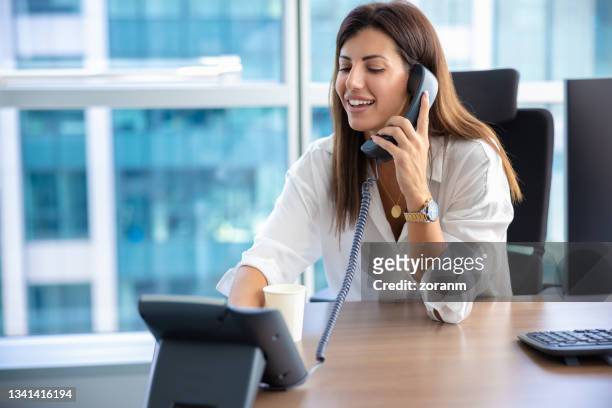businesswoman using landline and smiling at her desk - landline phone imagens e fotografias de stock