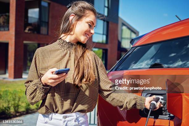 portrait of young woman charging electric car at front of her house - electric car home stock pictures, royalty-free photos & images