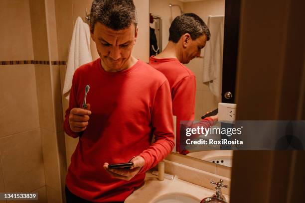 adult man brushing his teeth while looking at his cell phone in the bathroom. - brush teeth phone stock-fotos und bilder