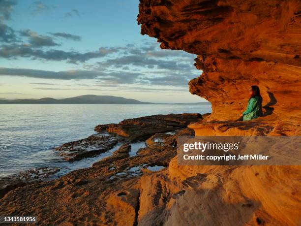 woman sitting along red sandstone cliff at sunset, lime bay, tasmania, australia - hobart stockfoto's en -beelden