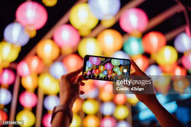 close up of woman's hands taking photos of illuminated and colourful traditional chinese lanterns with smartphone hanging on city street at night. traditional chinese culture, festival and celebration event theme - bunte festival night stock-fotos und bilder