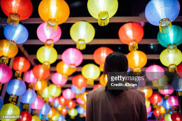 rear view of young asian woman looking up and admiring the hanging illuminated and colourful traditional chinese lanterns on city street at night. traditional chinese culture, festival and celebration event theme - asien metropole nachtleben stock-fotos und bilder