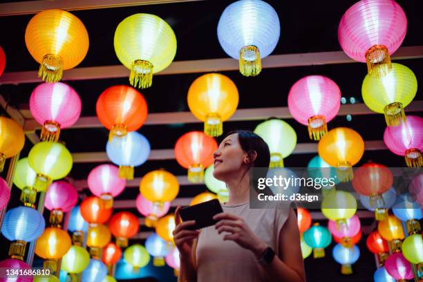 smiling young asian woman taking photos of illuminated and colourful traditional chinese lanterns with smartphone hanging on city street at night. traditional chinese culture, festival and celebration event theme - bunte festival night stock-fotos und bilder