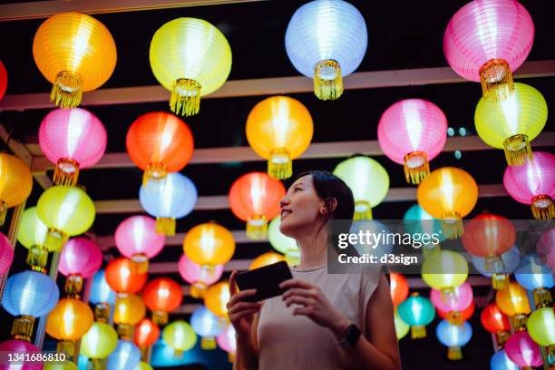 smiling young asian woman taking photos of illuminated and colourful traditional chinese lanterns with smartphone hanging on city street at night. traditional chinese culture, festival and celebration event theme - hong kong celebrates chinese new year stock pictures, royalty-free photos & images