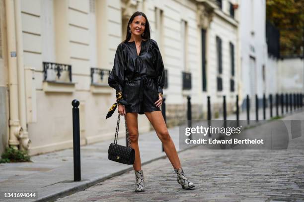 Alba Garavito Torre wears silver and orange rhinestones large pendants earrings, a black shiny leather oversized / puffy sleeves / belted short...