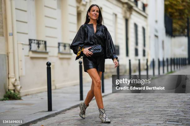 Alba Garavito Torre wears silver and orange rhinestones large pendants earrings, a black shiny leather oversized / puffy sleeves / belted short...