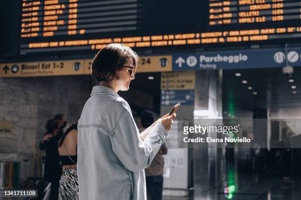 a young woman at a train station or airport looking at the arrival and departure boards - station de vacances fotografías e imágenes de stock