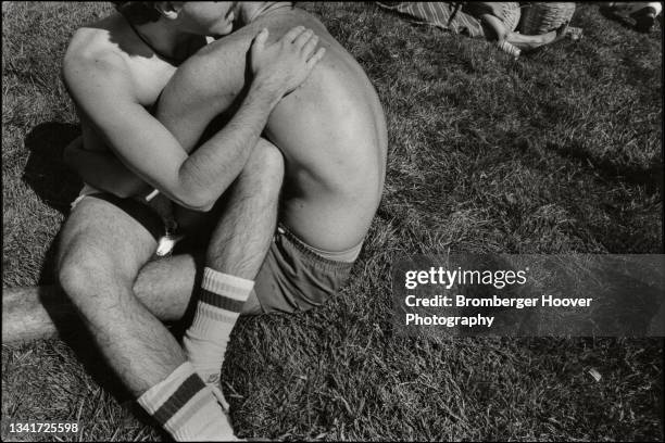 View of a two men, both shirtless, as they embrace, seated on the grass outside the Civic Center during the International Lesbian & Gay Freedom Day...