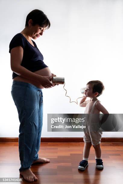 retrato de un niño de 2 años con su madre embarazada. el niño esta escuchando el vientre de su madre con un telefono hecho con unas latas de ojalata y una cuerda. fondo blanco. - niño fondo blanco stock pictures, royalty-free photos & images