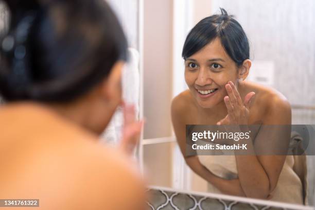 femme asiatique appliquant une crème pour la peau sur la joue dans la salle de bain - 薄ピンク photos et images de collection