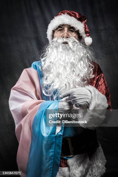 retro and desaturated santa claus on vintage gray background posing at camera, covered with the trnssexual flag, pointing at it with his hand. christmas, santa claus, diversity, lgbti, homosexuality and transsexuality concept. - gay santa claus stock pictures, royalty-free photos & images