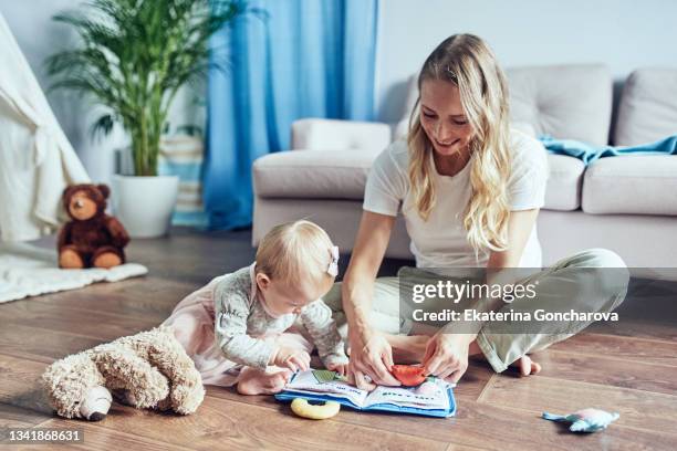 a babysitter with a 1-year-old girl is reading a children's playbook at home on the floor. - einjährig pflanzeneigenschaft stock-fotos und bilder