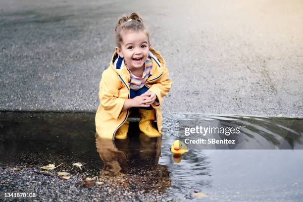 una niña de 3 años está agachada está jugando con un pato de goma en un charco - laarzen geel stockfoto's en -beelden