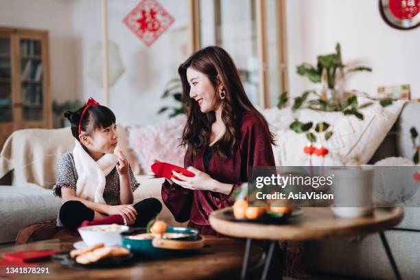 lovely daughter enjoying traditional snacks while helping her mother to prepare red envelops (lai see) at home for chinese new year - chinese kid stockfoto's en -beelden