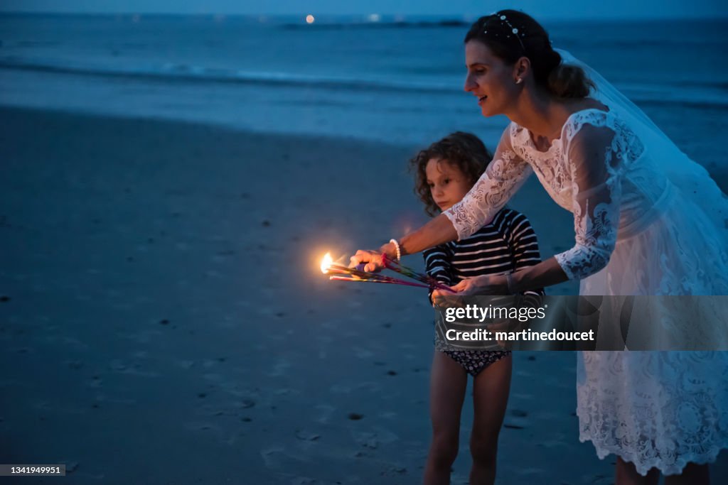 Mature bride lighting bengal fire with daughter on the beach at dusk.