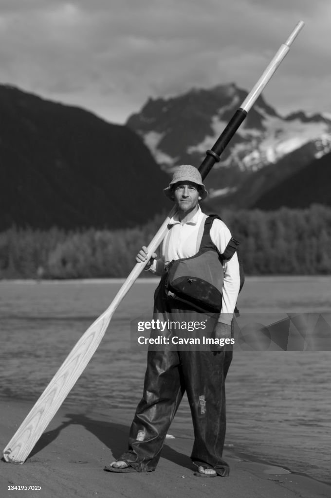 A man stands on the shores of a river for a portrait with an oar, wool hat and rain slicker overalls.